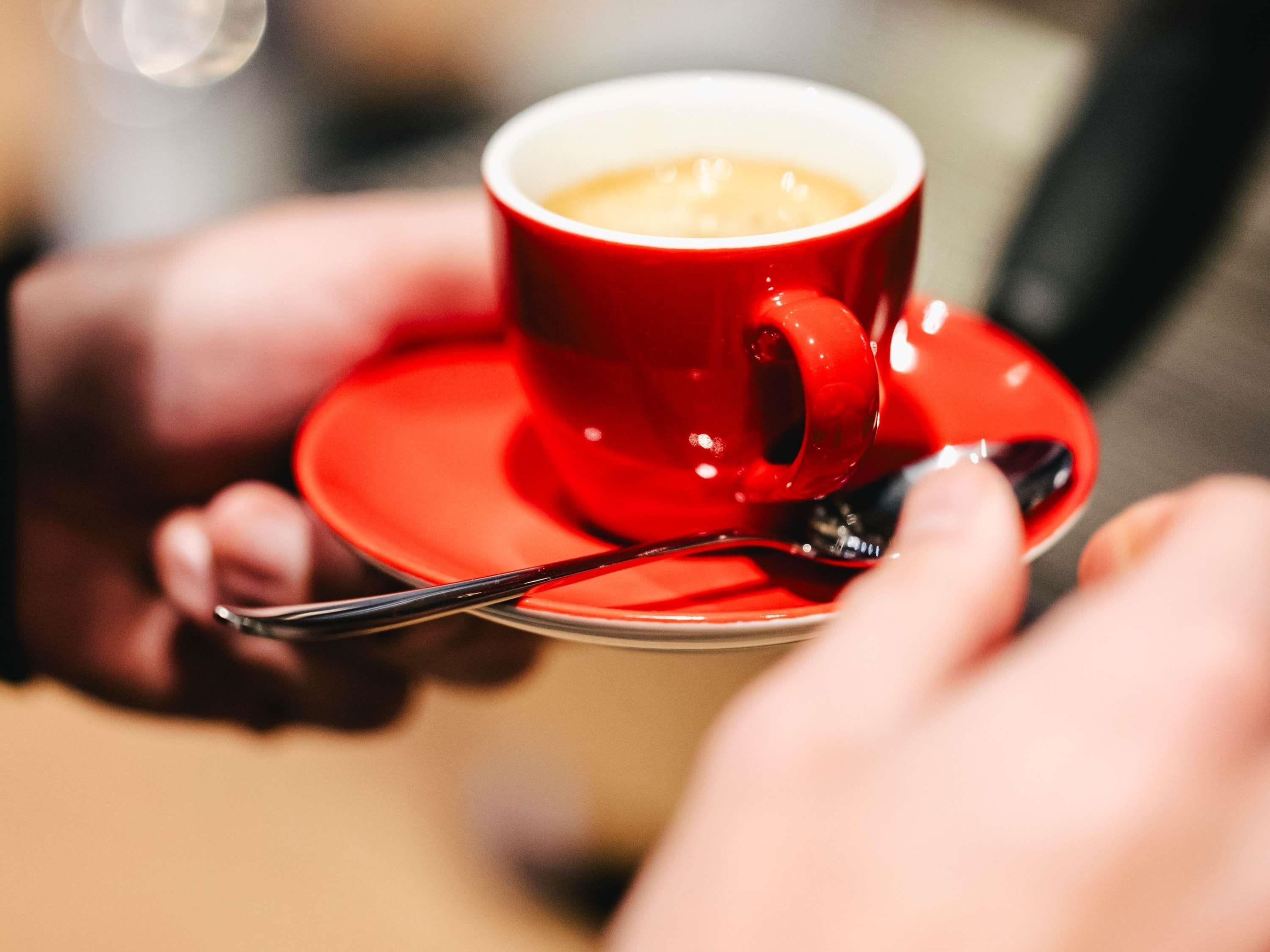 Una taza roja de café sobre un platito, con una cuchara, sostenida por una persona.