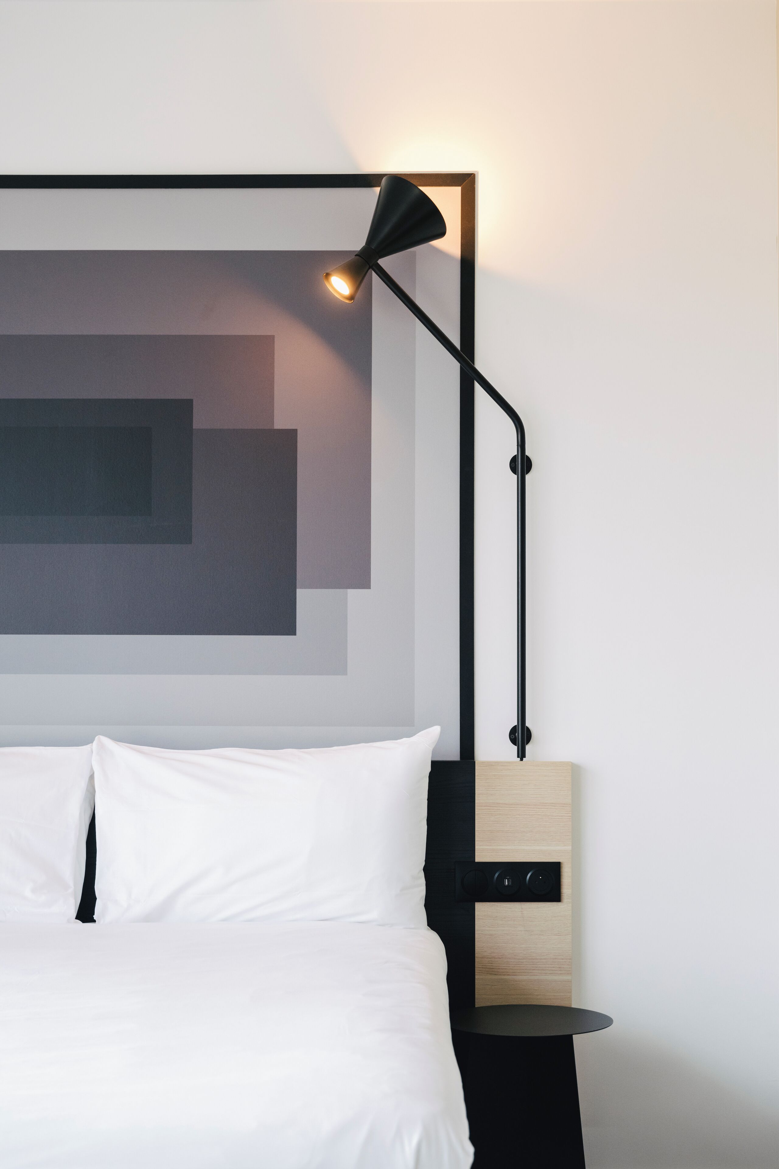 Un lit bien fait, avec du linge de lit blanc, une lampe de sol noire au design moderne et une œuvre d’art murale minimaliste.