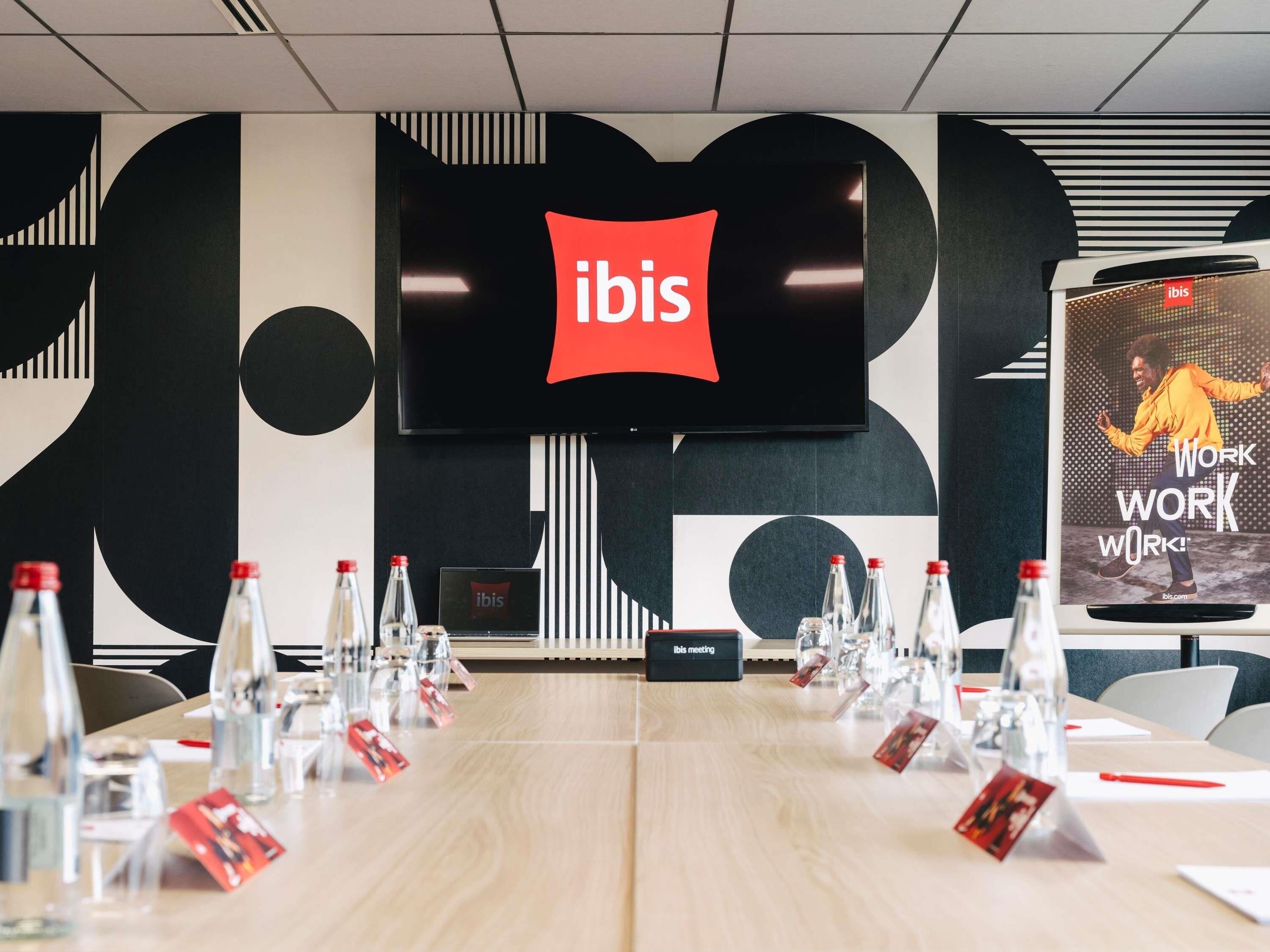 A conference room with a large screen displaying the ibis logo, a meeting table with name cards, and water bottles.