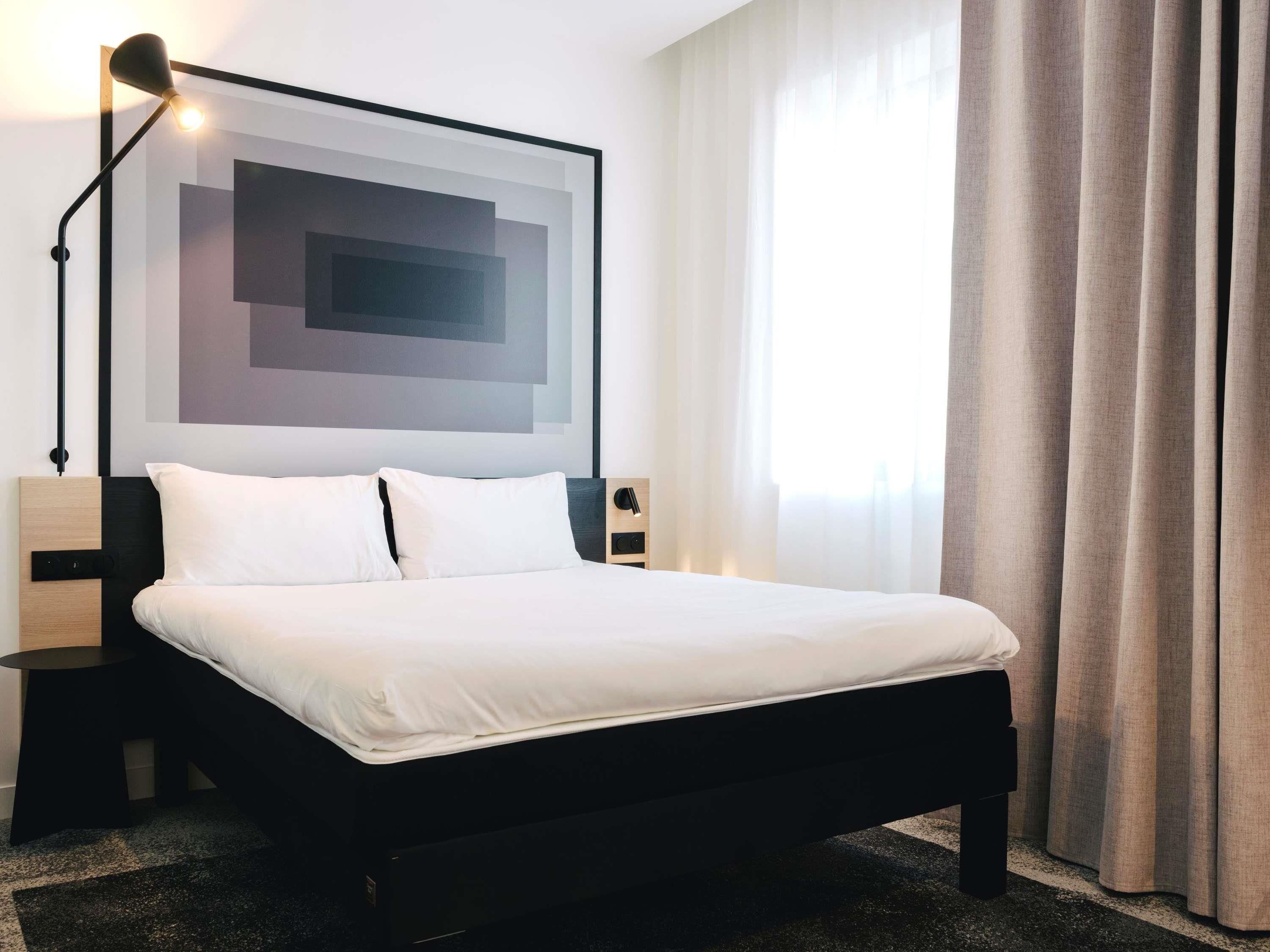 A hotel room with a bed, a nightstand, a lamp, a wall art piece, and a window with curtains.