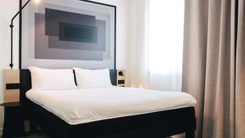 A hotel room with a bed, a nightstand, a lamp, a wall art piece, and a window with curtains.