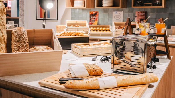 Mostrador de panadería con panes y pasteles diversos, un cortador de pan y una estación de café.