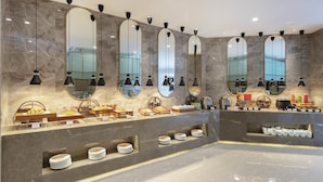 Un moderno vestíbulo de hotel con paredes de mármol, múltiples espejos y una zona de bufé con diferentes alimentos y platos.