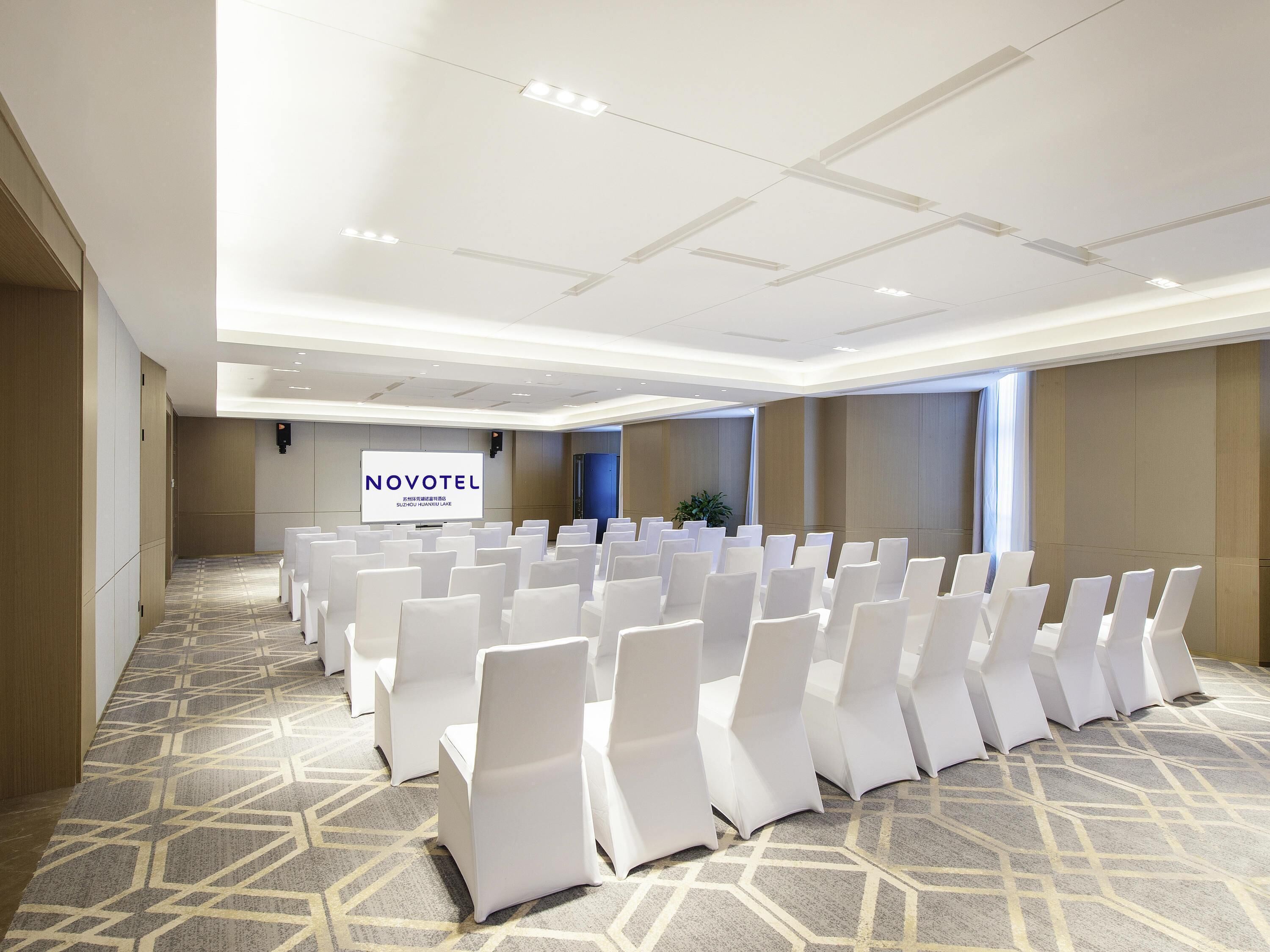 A conference room with rows of white chairs, a podium, and a large screen displaying 'NOVOTEL'.
