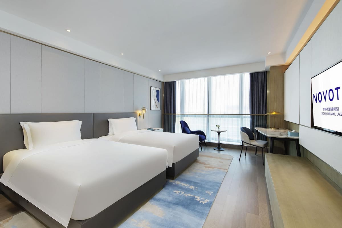 Een hotelkamer met twee bedden, een bureau, stoelen, een televisiescherm en een groot raam.