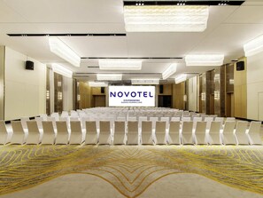 Une salle de conférence avec des rangées de chaises blanches, un grand écran affichant le mot « NOVOTEL » et un tapis à motifs.