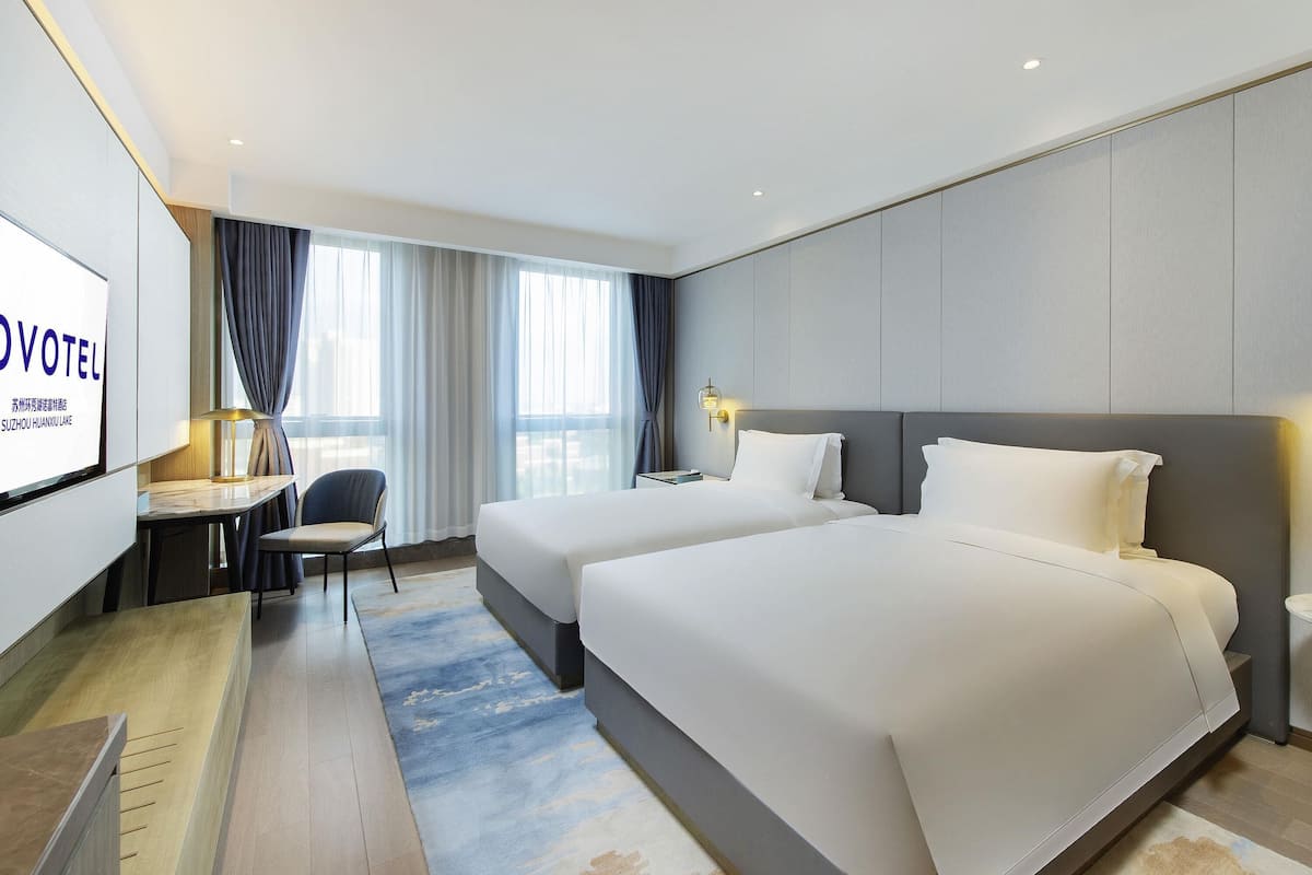 Hotelkamer met twee bedden, een bureau, een stoel, een televisie en een groot raam met gordijnen.