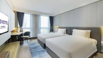 A hotel room with two beds, a desk, a chair, a TV, and a large window with curtains.