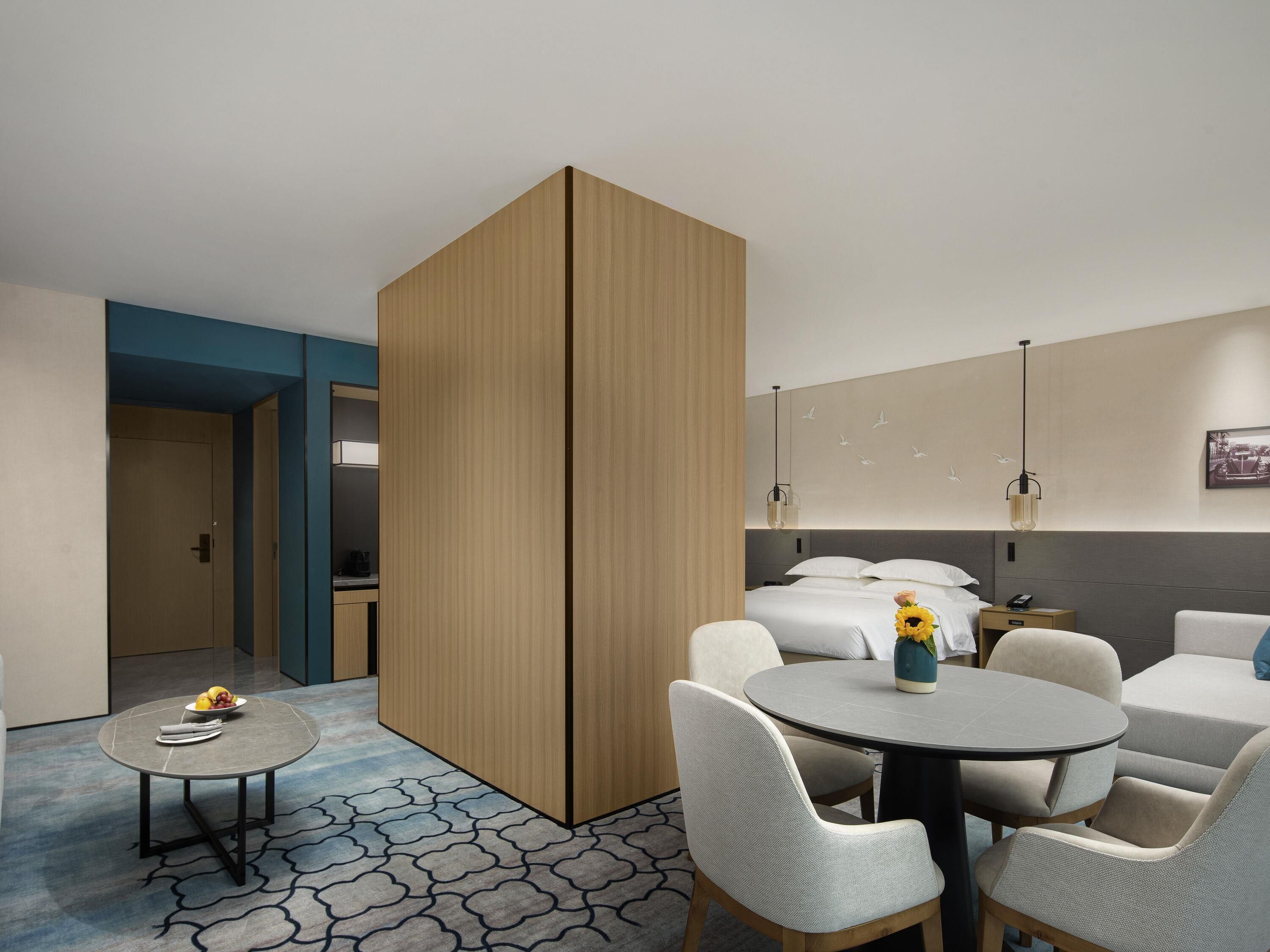 A modern hotel room with a round dining table, a sofa, a separate sleeping area, and a bathroom visible through an open door.