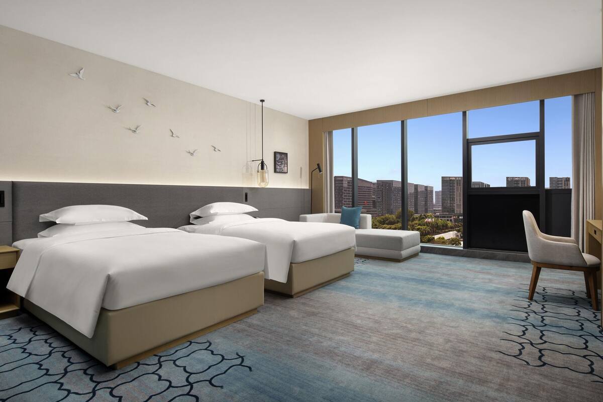 Quarto de hotel com duas camas, uma janela grande com vista para a cidade, uma escrivaninha e uma cadeira.