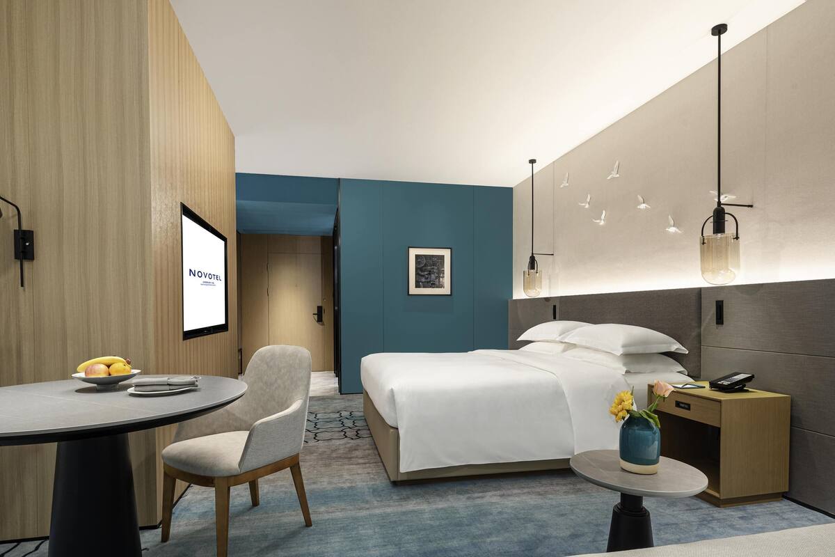 Quarto de hotel moderno com cama, escrivaninha, cadeira, mesinha de centro com um vaso e TV fixada na parede.
