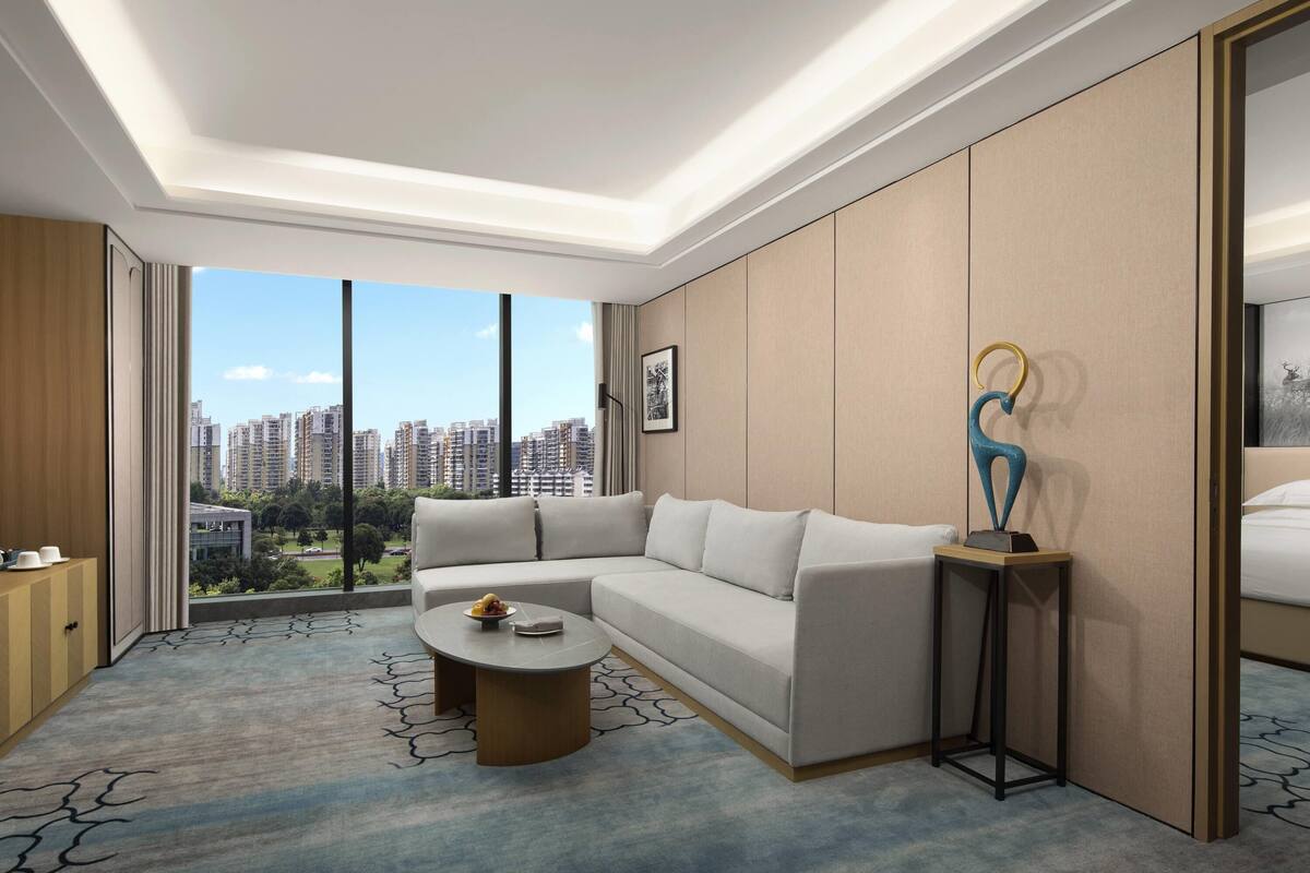 Quarto de hotel moderno com uma janela grande, um sofá, uma mesa de centro e uma escultura decorativa.