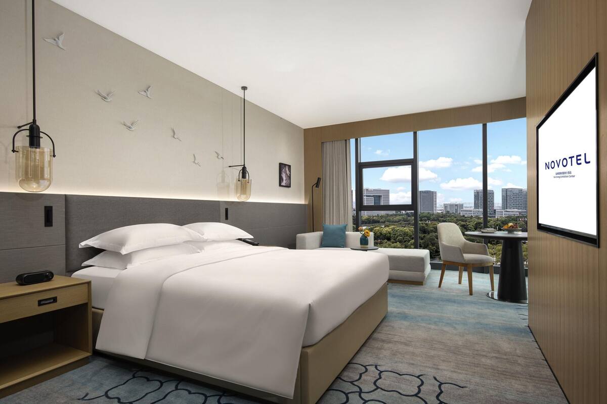 Quarto de hotel moderno com uma cama grande, mesas de cabeceira, escrivaninha e vista para a cidade.
