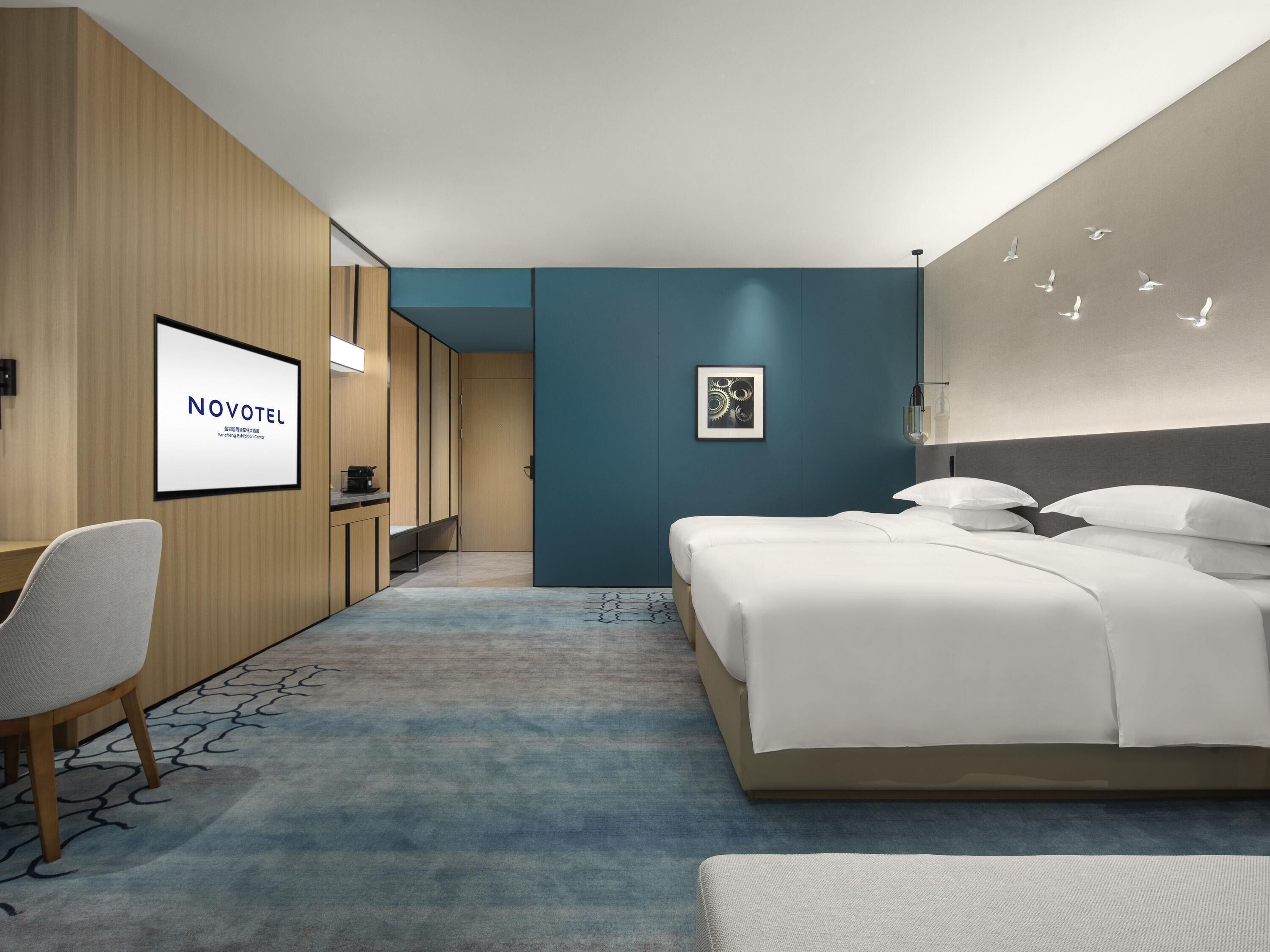 Una habitación de hotel con una cama, un escritorio, una silla, un cuadro enmarcado y una lámpara fijada a la pared.