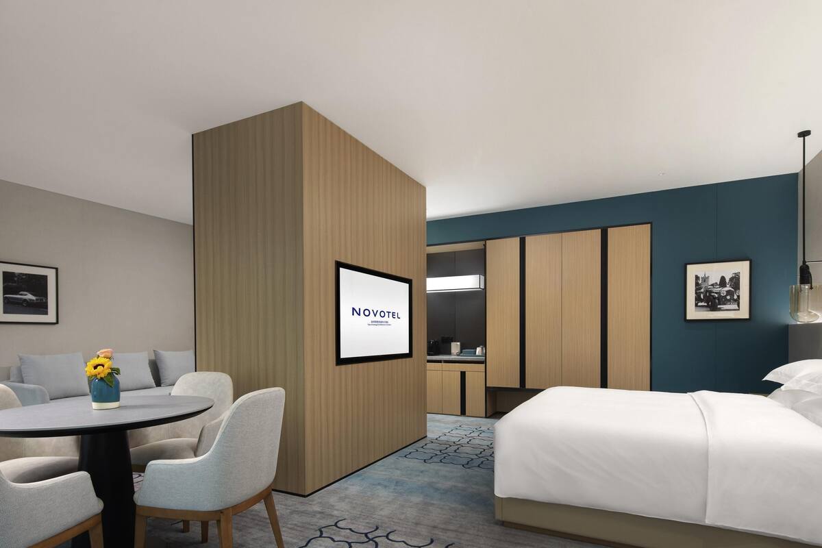 Quarto de hotel com cama, escrivaninha com cadeiras, mesinha com um vaso, cozinha compacta e uma moldura na parede.