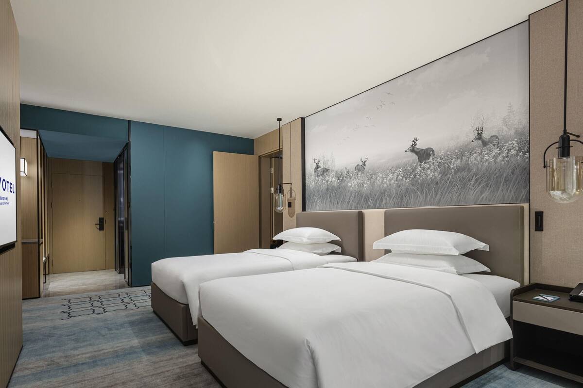 Quarto de hotel com duas camas, uma obra de arte grande pendurada na parede, uma mesa de cabeceira com luminária e uma porta que dá para outro quarto.