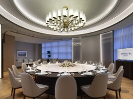 Un salón de conferencias con una mesa redonda, sillas blancas, una lámpara de araña y una pantalla de televisión que muestra 'NOVOTEL'.