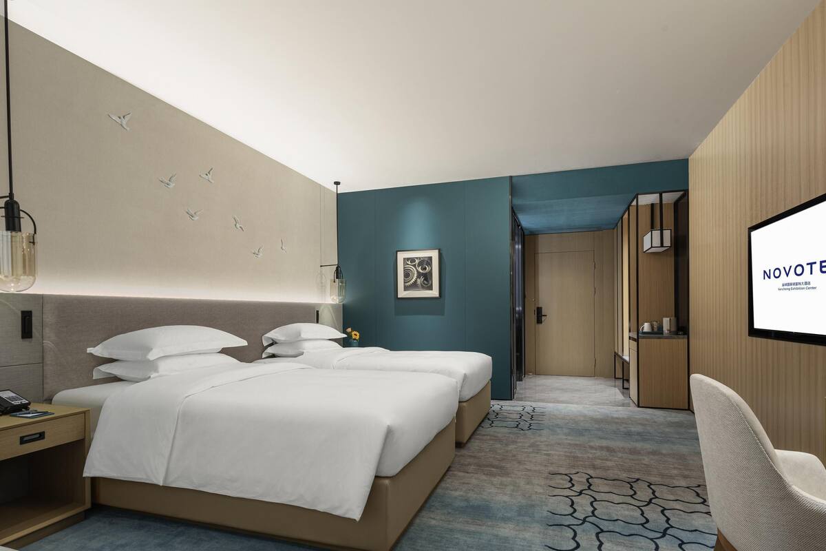 Quarto de hotel moderno com uma cama grande, uma escrivaninha, uma cadeira e uma tela de TV exibindo 'NOVOTEL'.