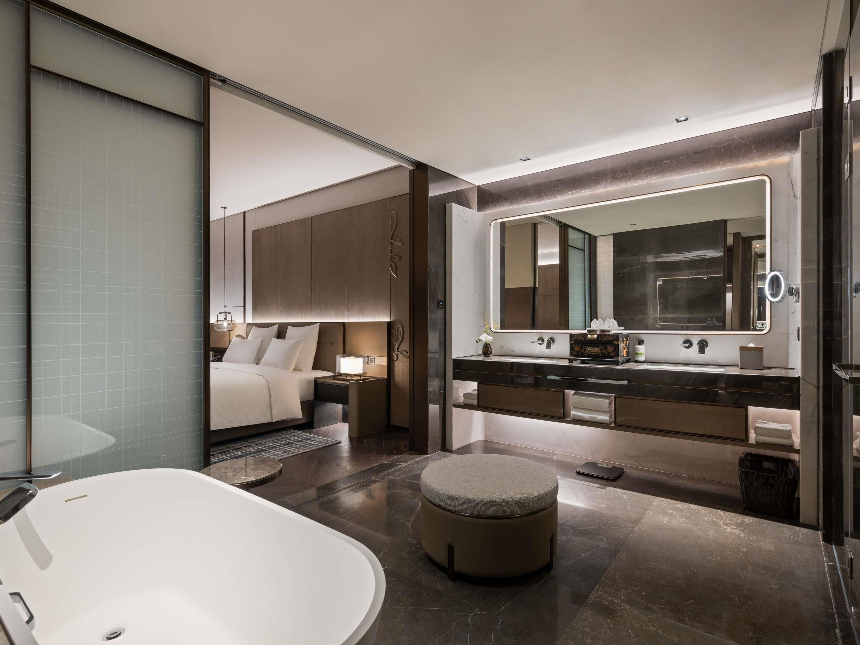 A modern bathroom with a large bathtub, a round vanity with a mirror, and a separate bedroom area with a bed and nightstands.