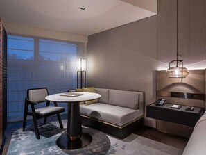 Habitación de hotel moderna con una mesa redonda, dos sillas, un sofá, una mesita de noche con lámpara y vistas a la ciudad a través de amplias ventanas.