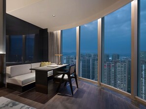 Una sala de estar moderna con un sofá, un escritorio y una silla, ventanas amplias con vista a la ciudad y un techo curvo.