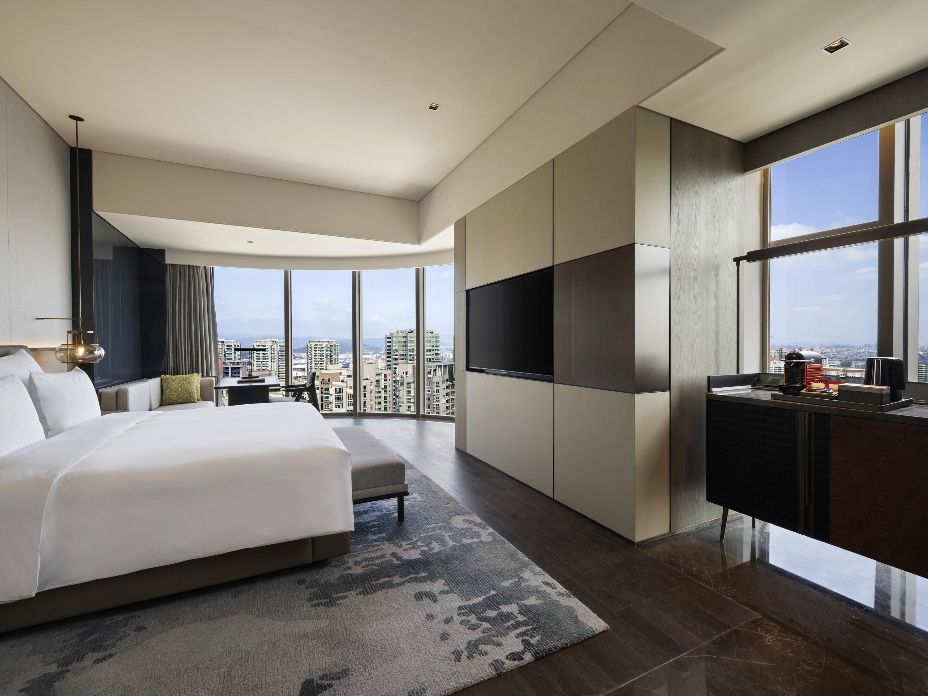 Quarto de hotel moderno com uma cama grande, televisão de tela plana, escrivaninha com cafeteira e vista para a cidade.