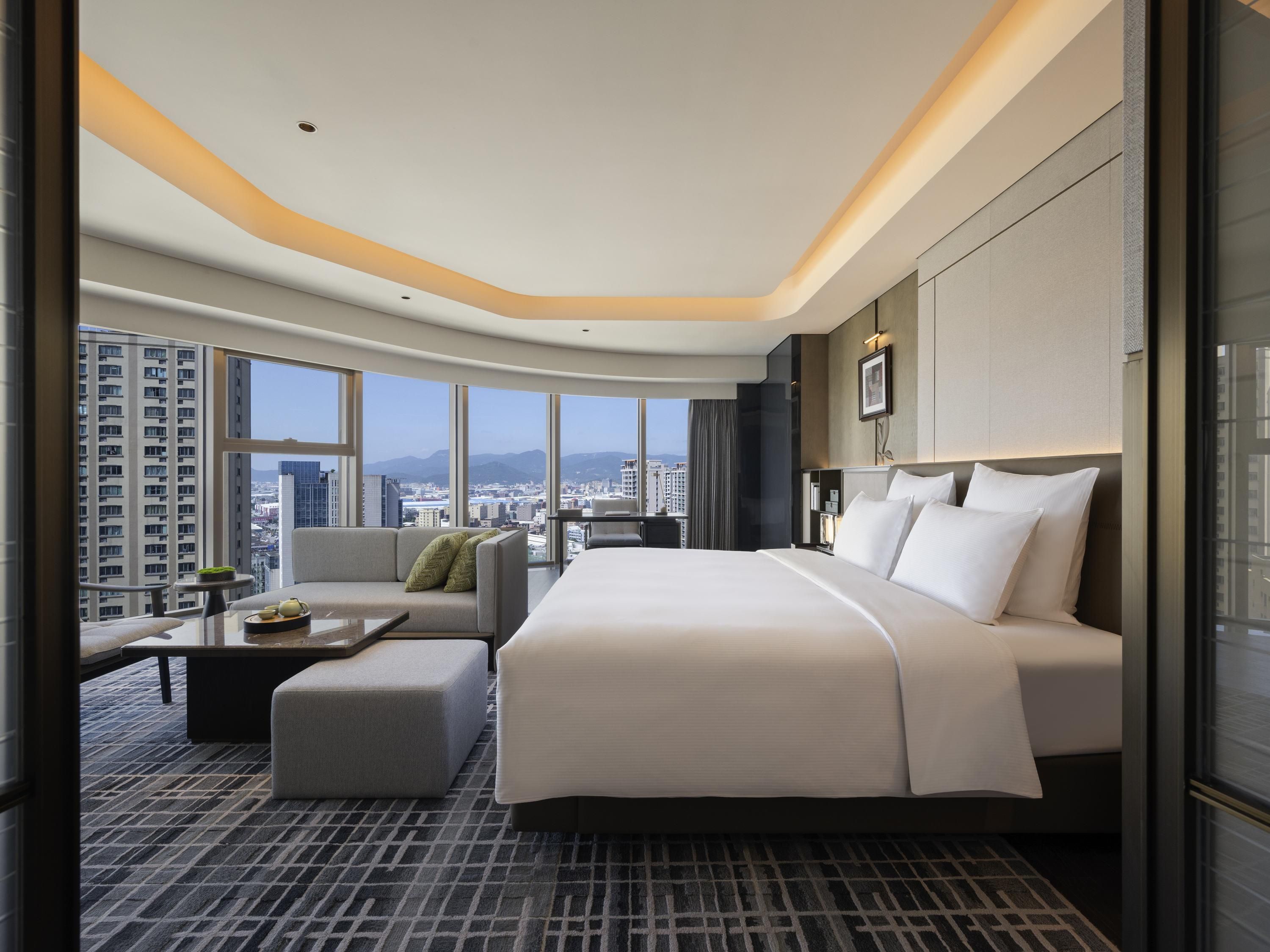 Quarto de hotel com uma cama grande, um sofá, uma mesa pequena com bandeja e vista para a cidade através de janelas amplas.