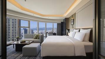 Quarto de hotel com uma cama grande, um sofá, uma mesa pequena com bandeja e vista para a cidade através de janelas amplas.