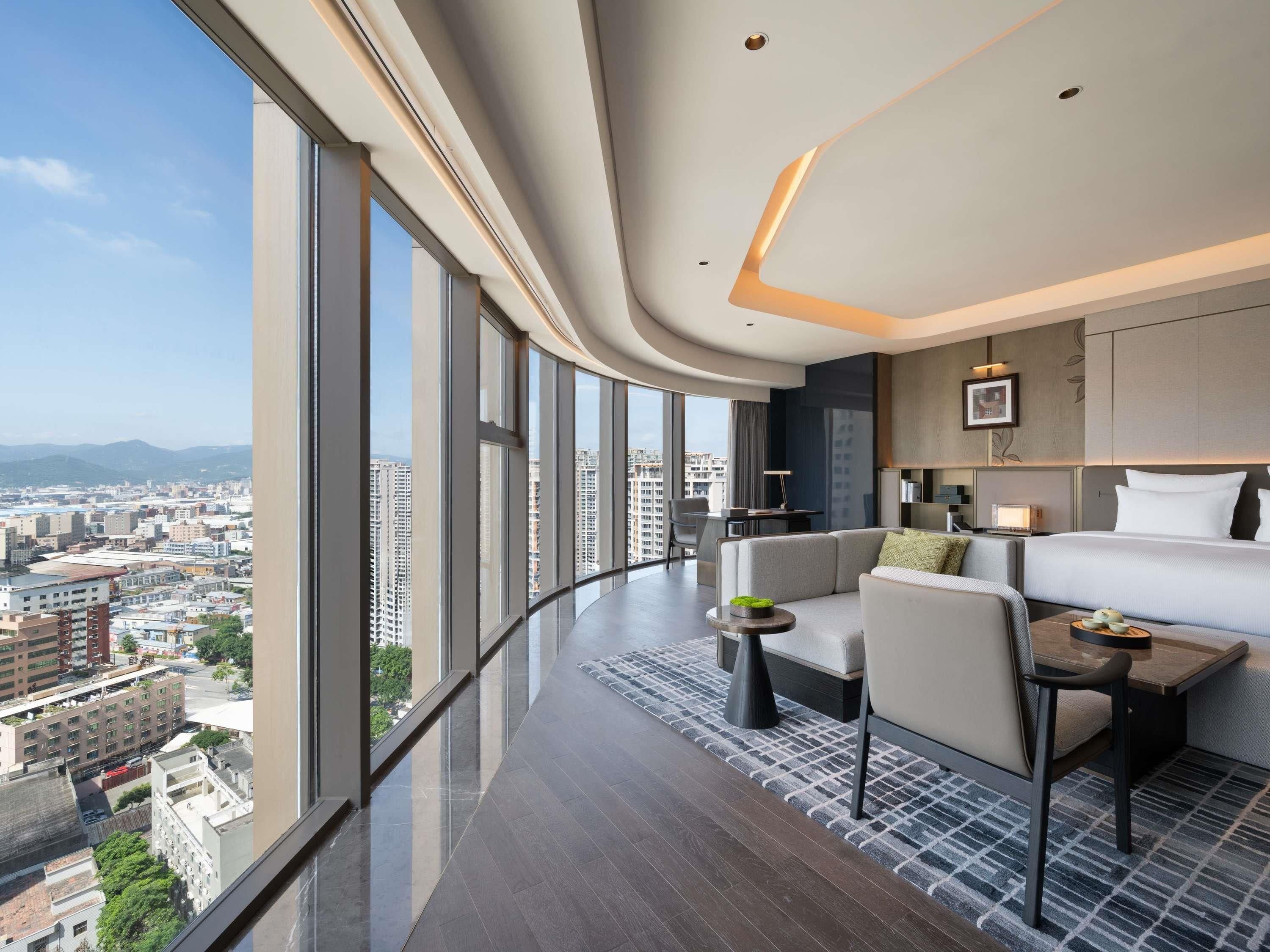 A modern hotel room with a curved ceiling, large windows offering a city view, a sofa, a dining table, and a bed.