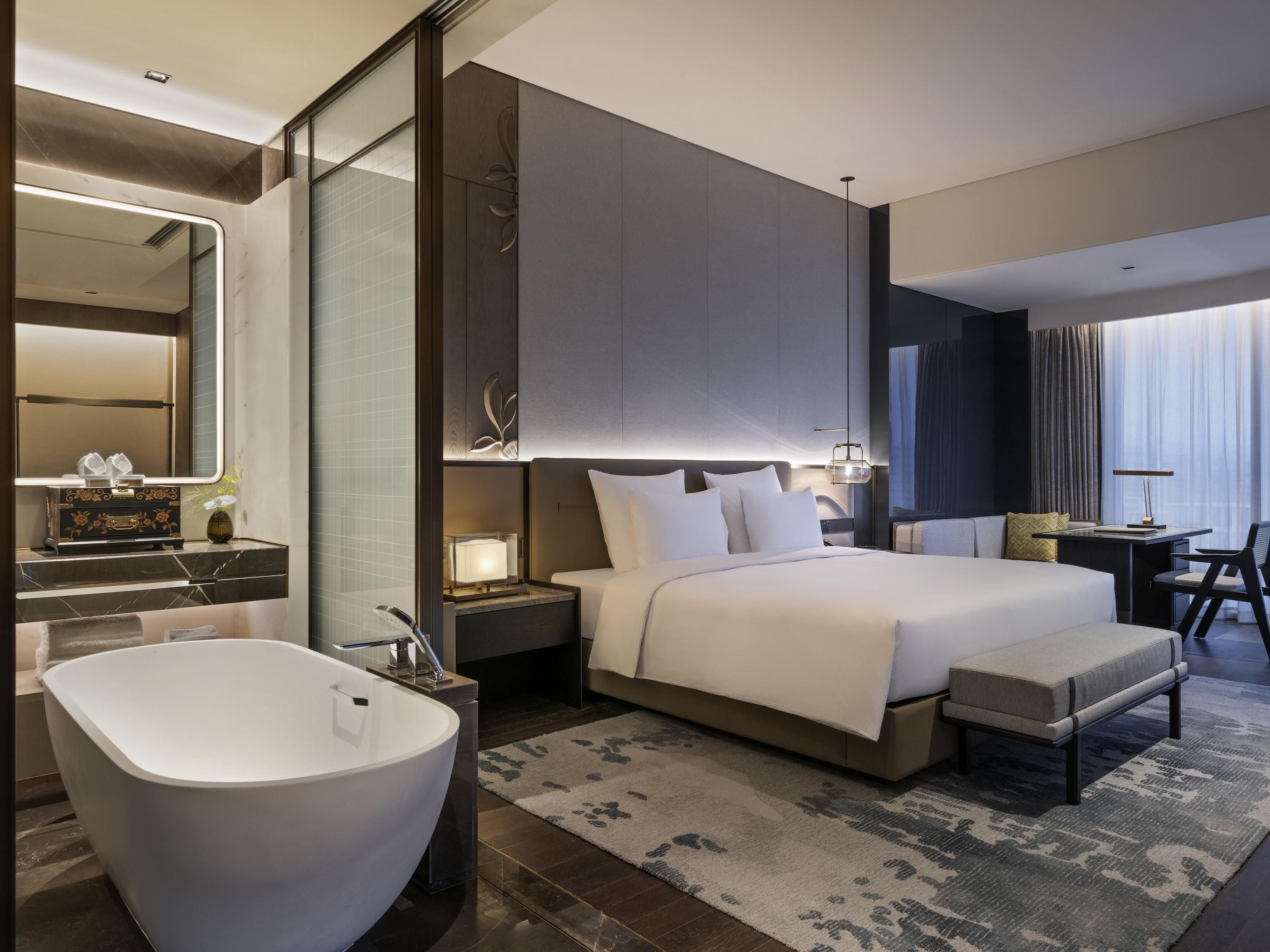 A modern hotel room with a large bed, a separate sitting area, a large bathroom with a freestanding bathtub, and a large mirror.