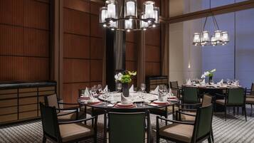 A well-lit dining area with round tables, green upholstered chairs, and a large window in the background.