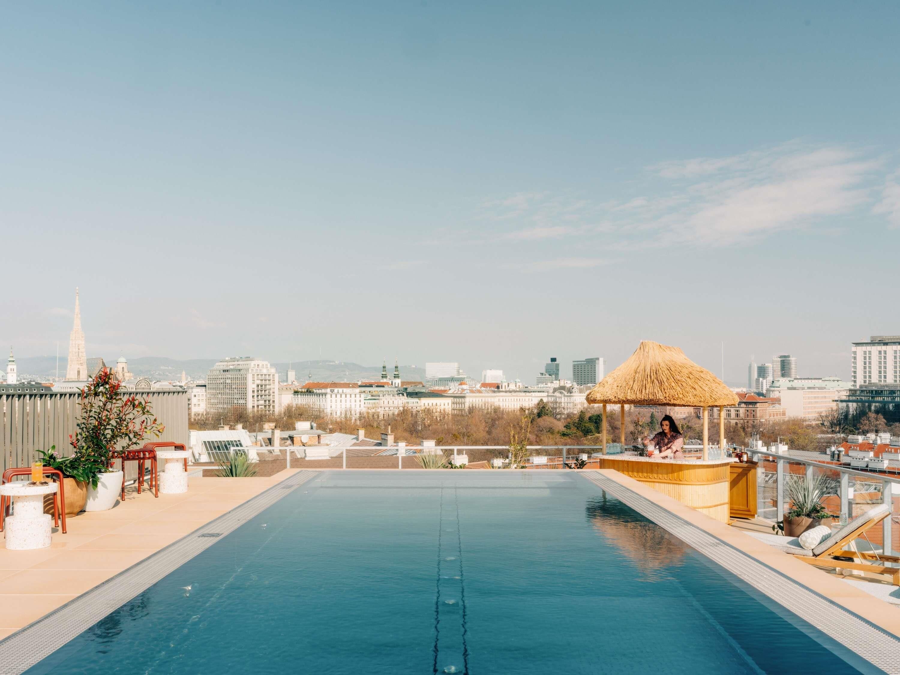 A rooftop pool with a cityscape view, a thatched umbrella, and lounge chairs.