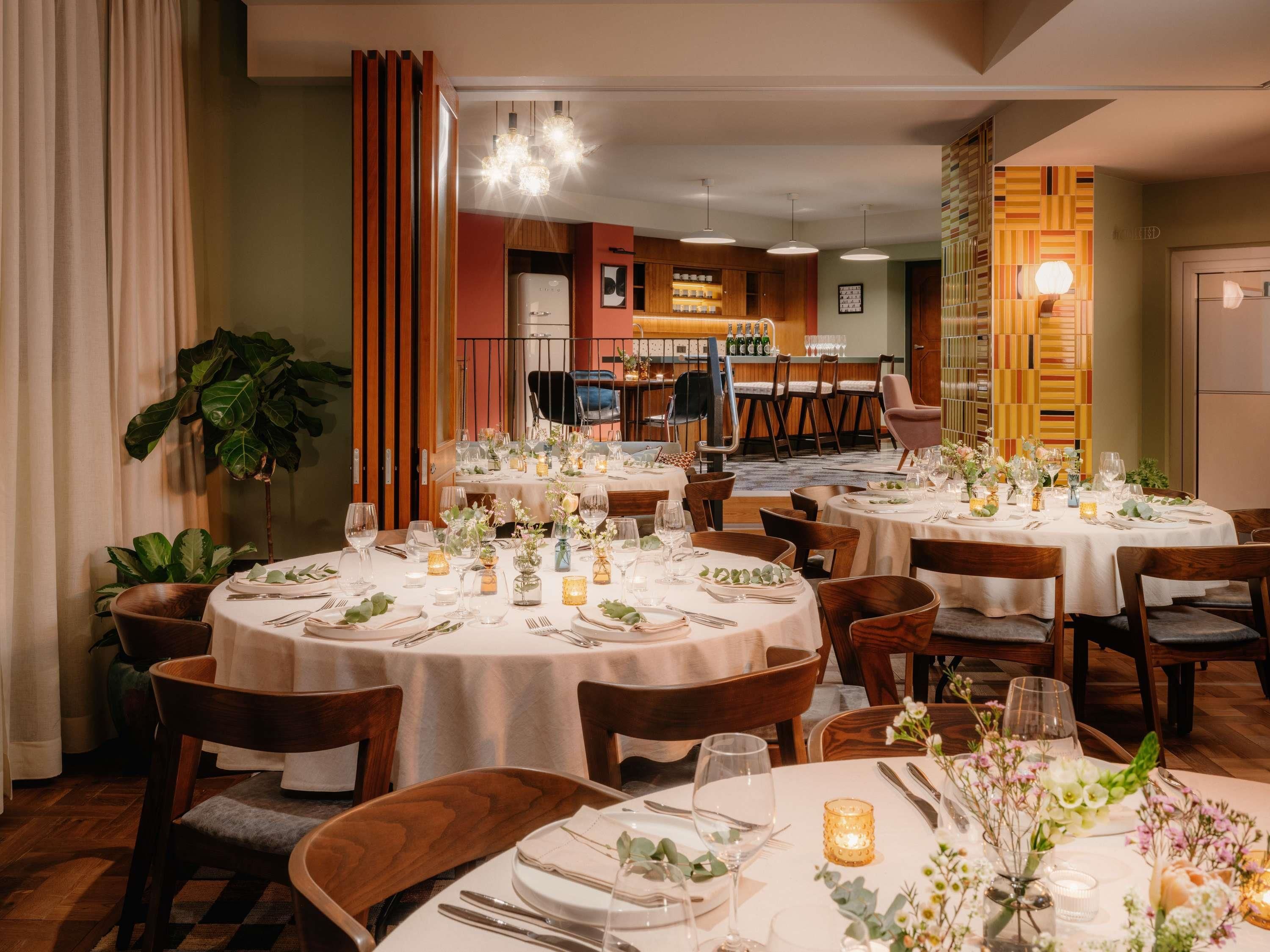 A dining area with round tables set for a meal, chairs, and a bar area in the background.