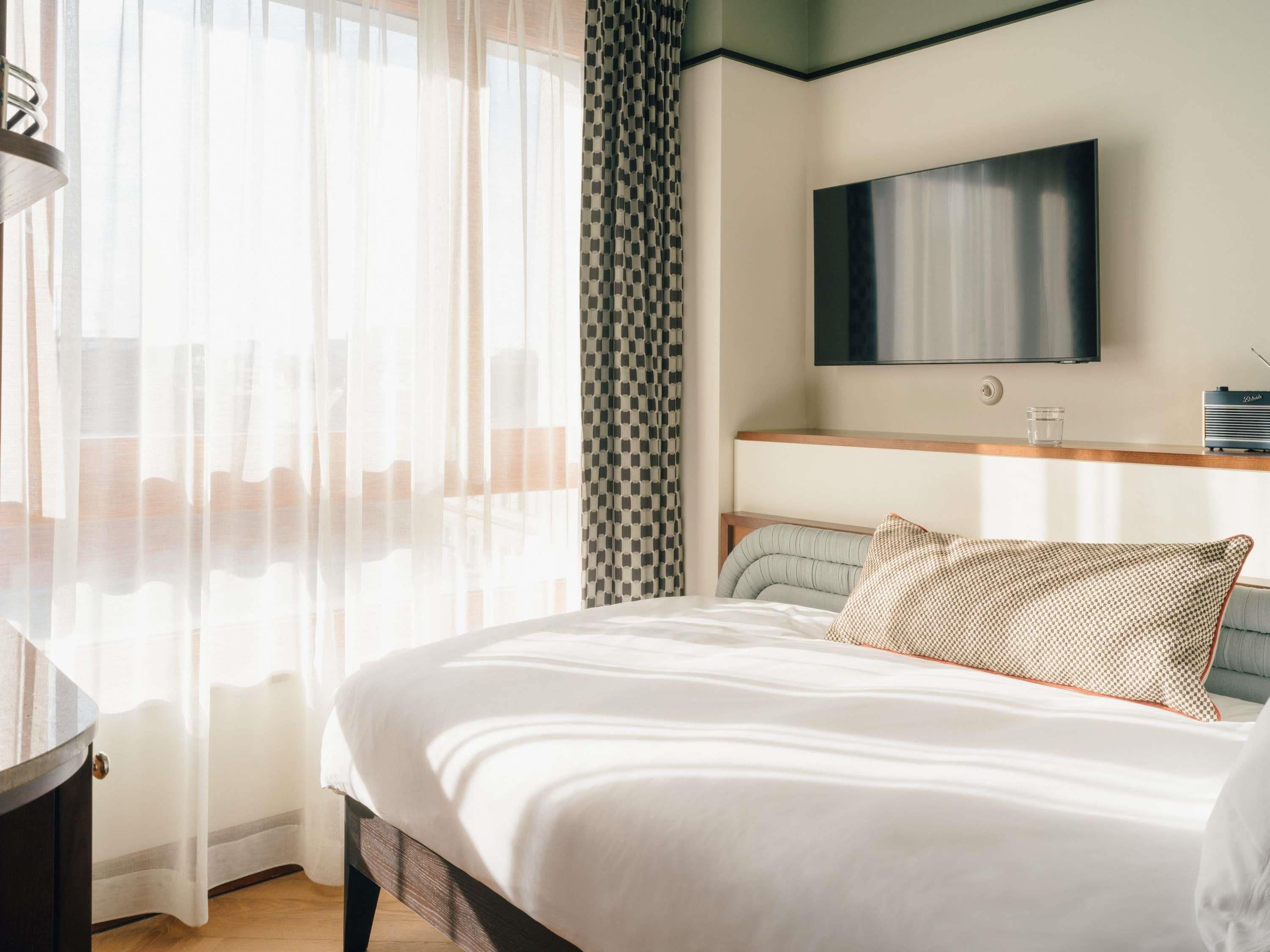 Una habitación de hotel moderna con una cama grande, televisión montada en la pared y una ventana con cortinas traslúcidas.