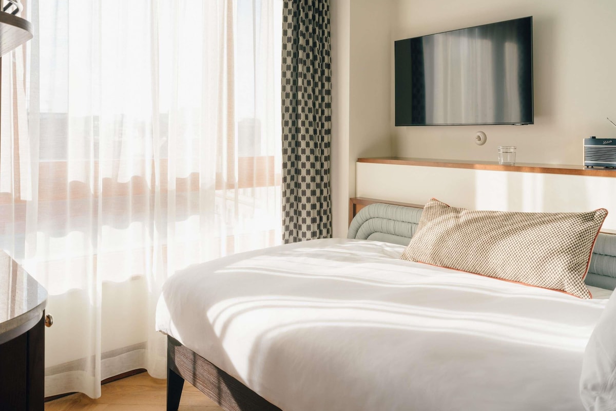 A modern hotel room with a large bed, a television mounted on the wall, and a window with sheer curtains.