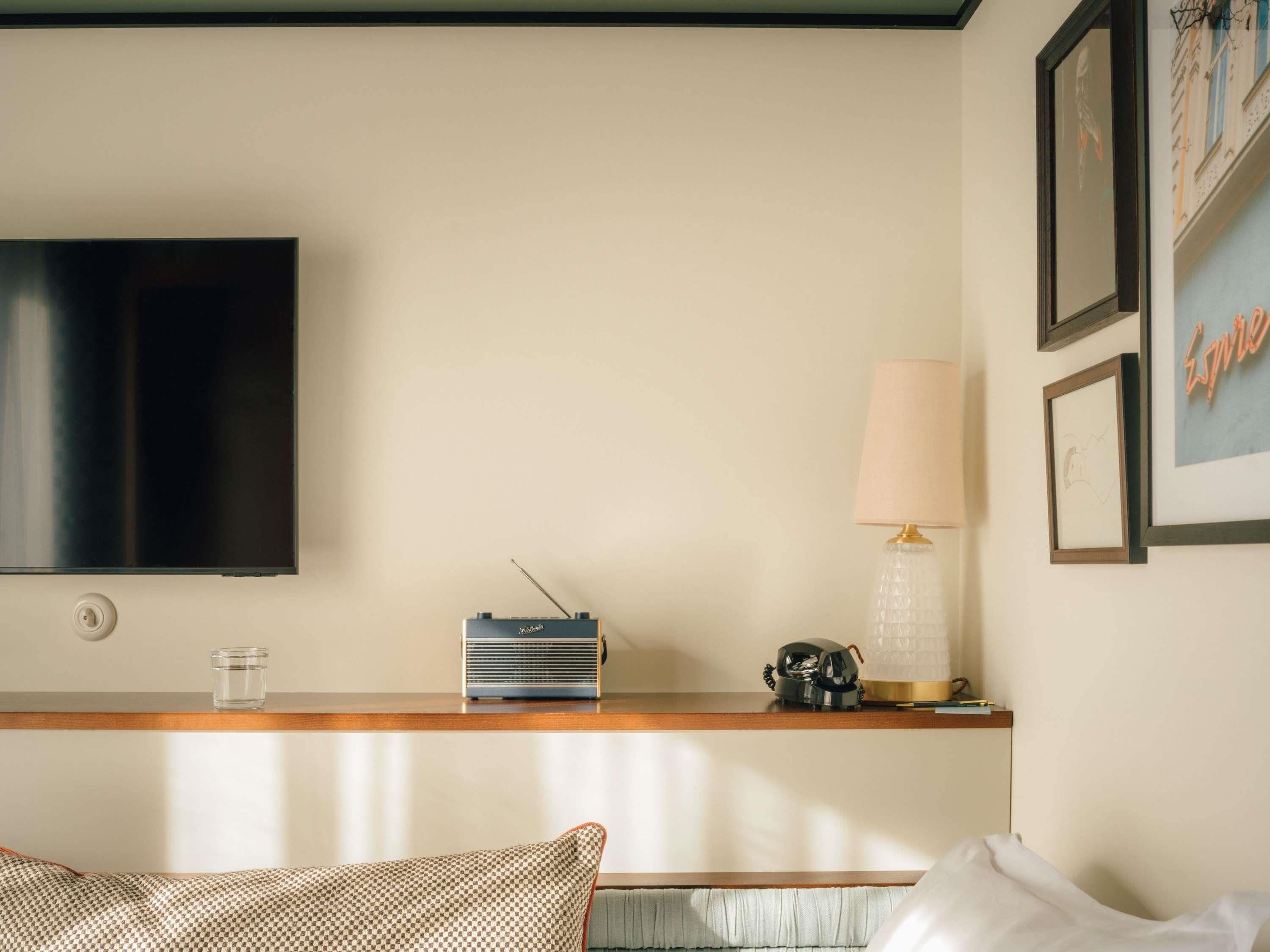 A hotel room with a flat-screen TV mounted on the wall, a radio on a wooden console, a lamp, and framed artwork.