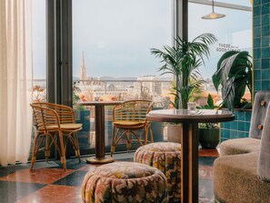 A modern interior with wicker chairs, a round table, and a view of a cityscape through large windows. - The Hoxton, Vienna (Vienna)