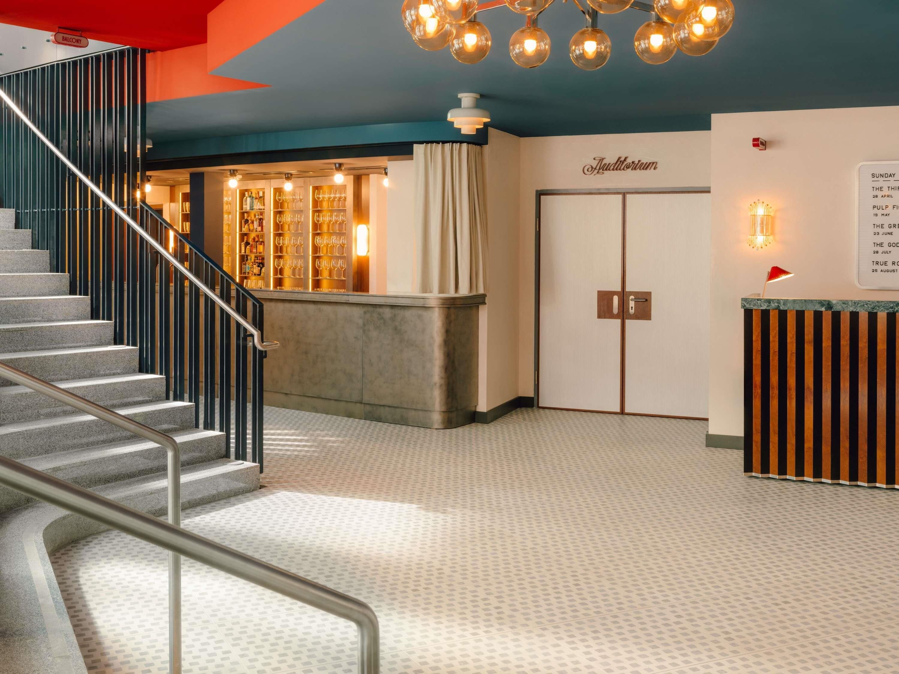 Um saguão de hotel com uma escadaria, uma mesa de recepção, uma parede com a placa de um auditório e um mural com anúncios de eventos.
