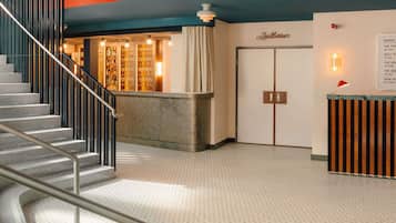 Um saguão de hotel com uma escadaria, uma mesa de recepção, uma parede com a placa de um auditório e um mural com anúncios de eventos.