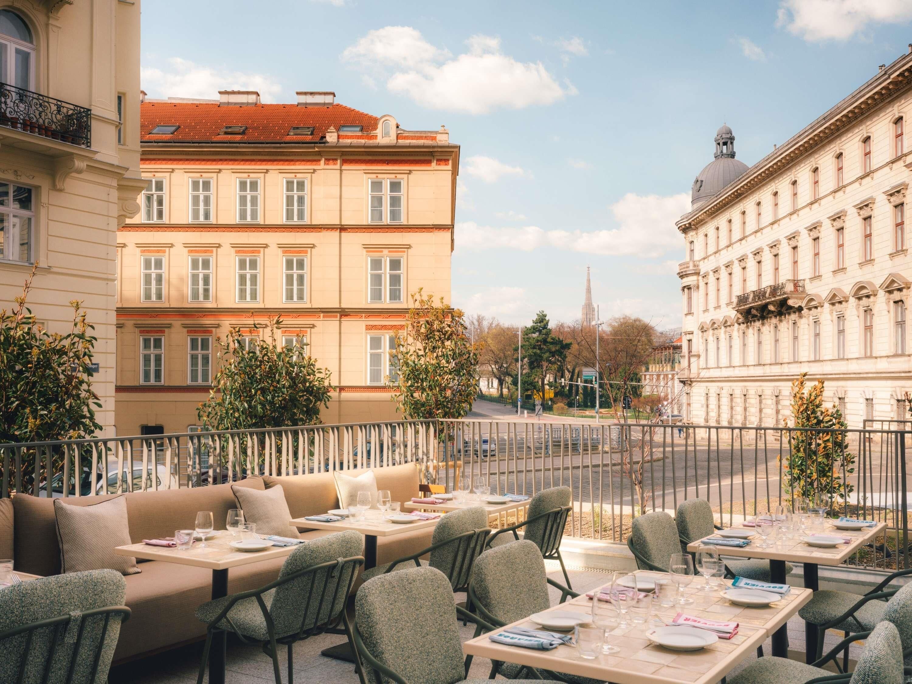 Un café en plein air, avec des tables et des chaises, entouré d’une clôture métallique, et des bâtiments en arrière-plan.