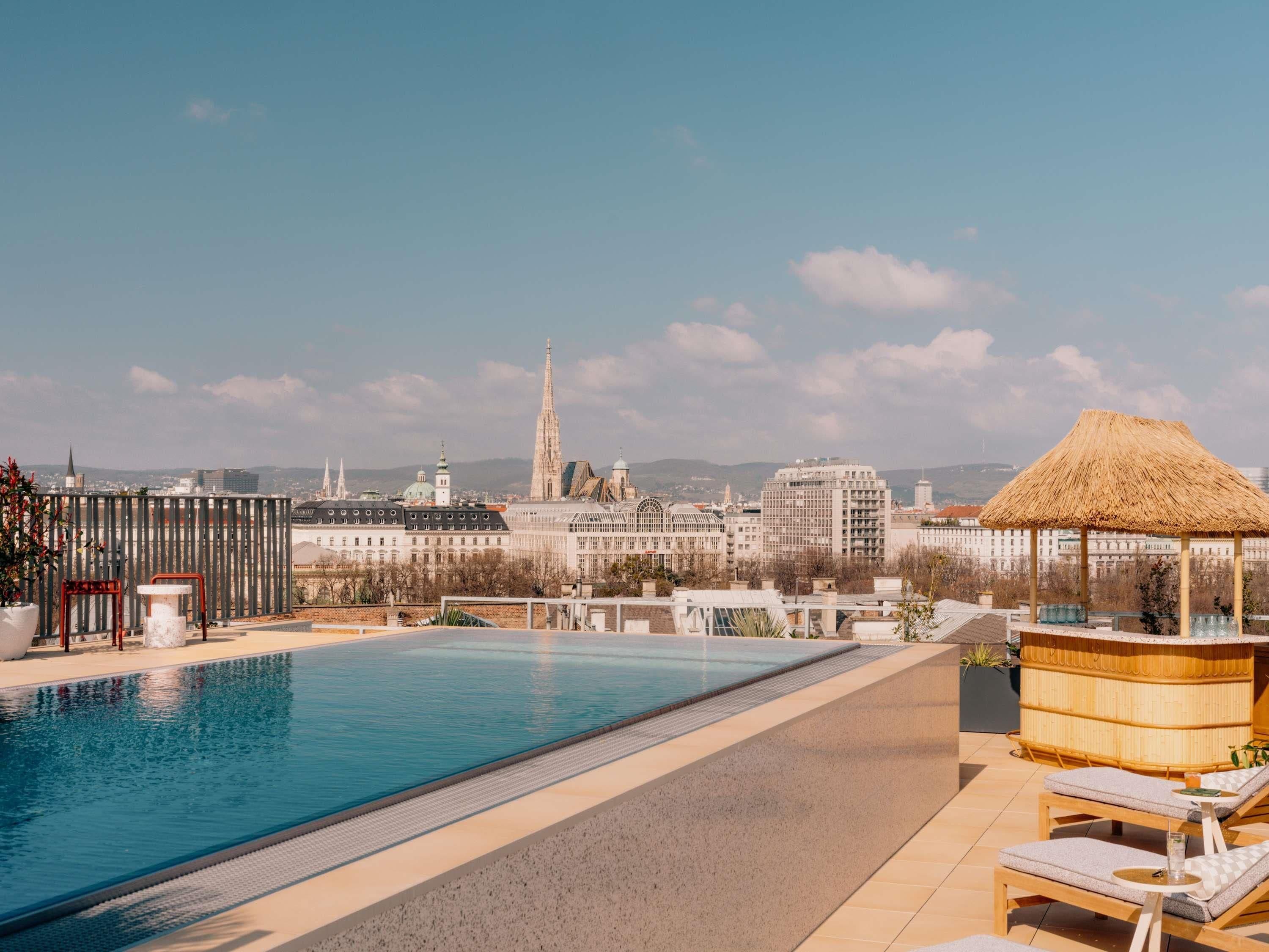 Piscina no terraço com vista para a paisagem urbana, cadeiras de descanso e uma estrutura com telhado de palha.