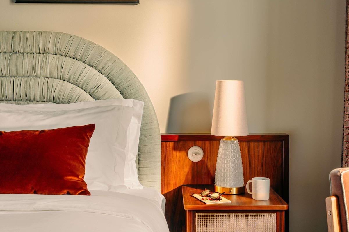 A neatly made bed with pillows, a bedside lamp, a small table with a cup and glasses, and framed artwork on the wall.
