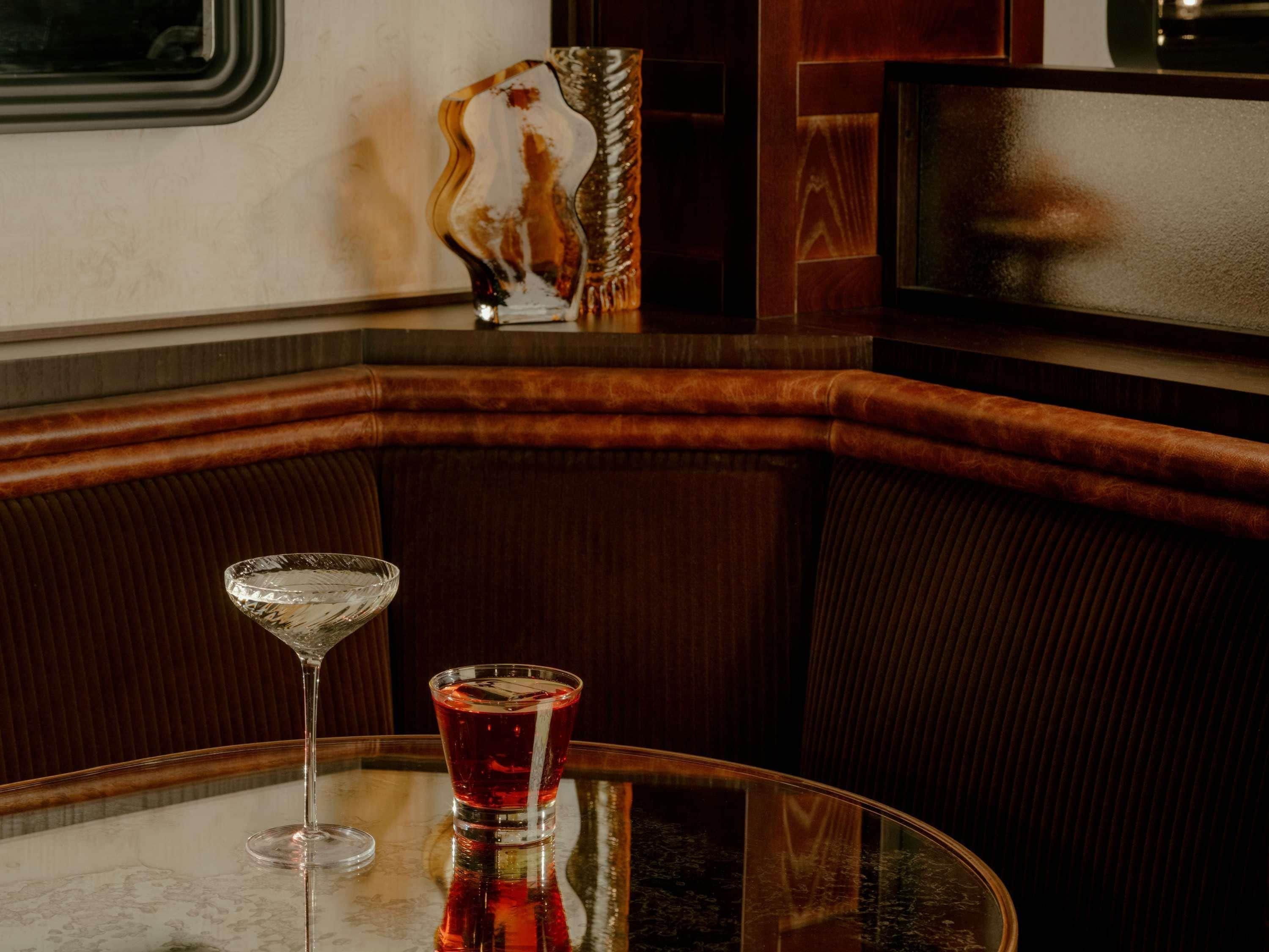 A table with a glass of red beverage and a clear glass, set in a corner with a cushioned bench.