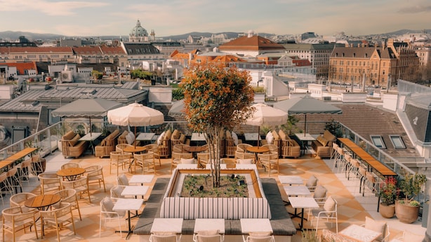 An outdoor terrace with seating, tables, and umbrellas, overlooking a cityscape with historic buildings. - The Hoxton, Vienna (Vienna)