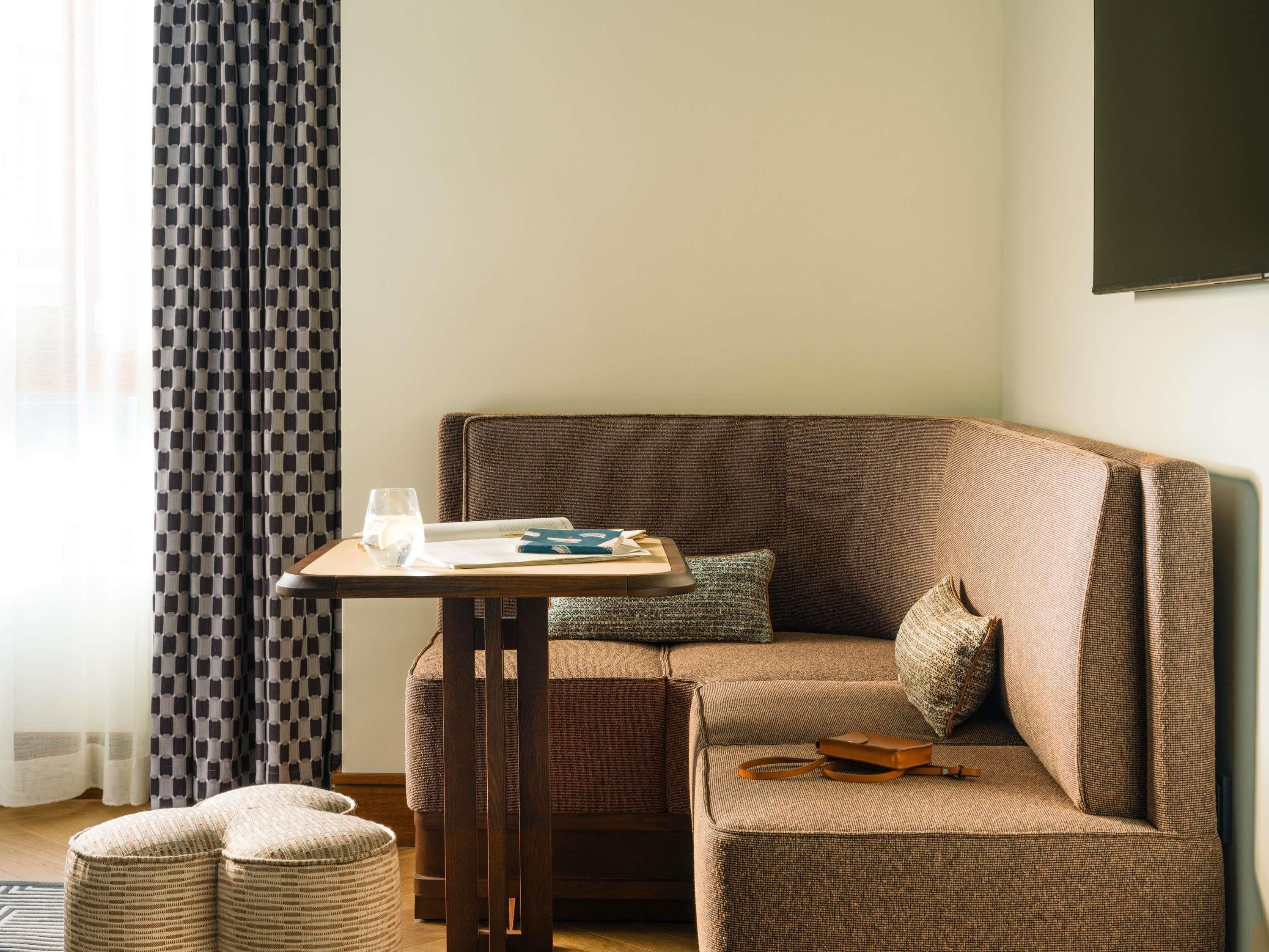 A cozy room with a brown sofa, a small wooden table with a glass of water and a book, and a patterned curtain.