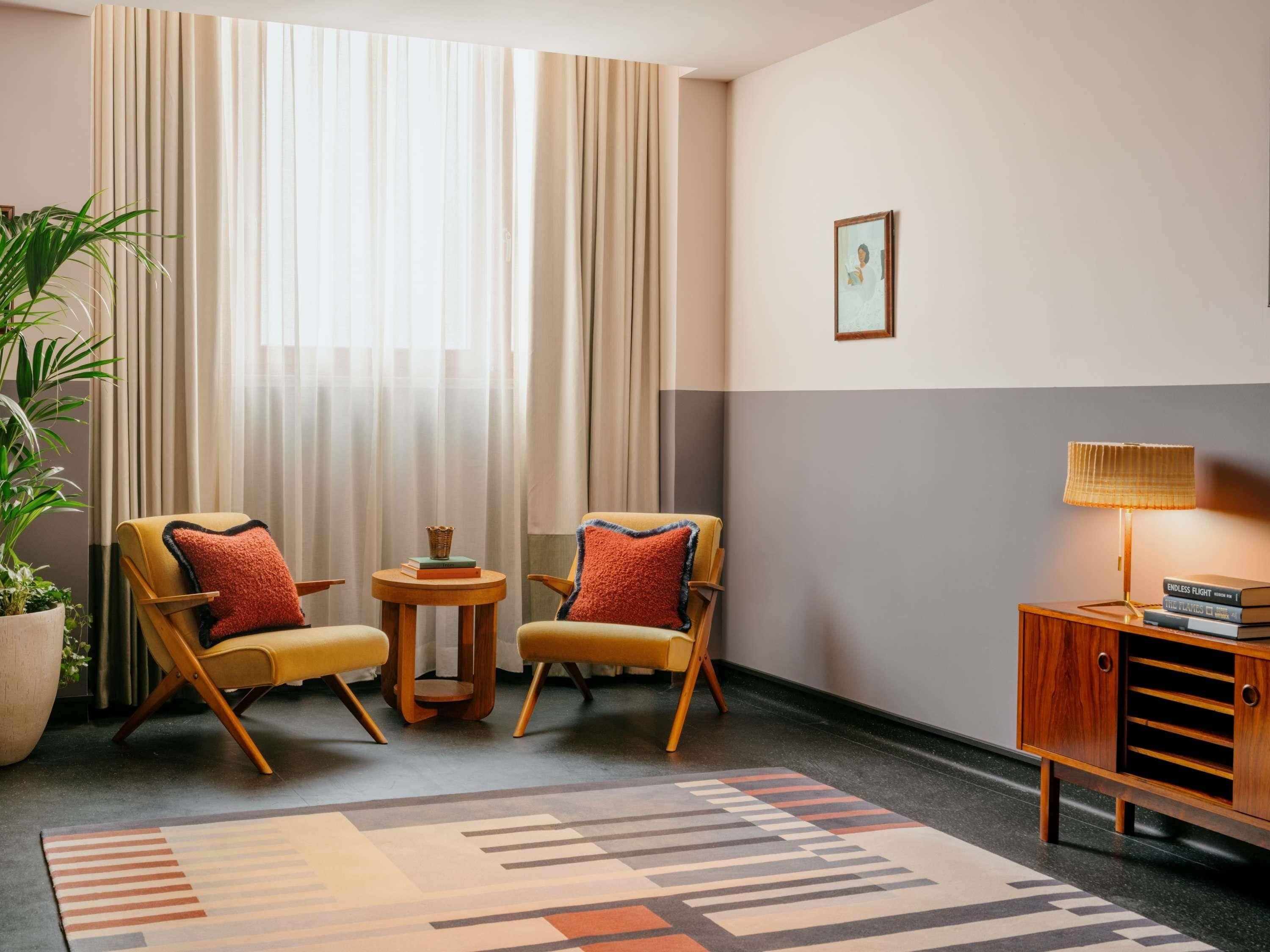 Habitación de hotel con dos sillones, una mesita, una lámpara, una estantería y un amplio ventanal con cortinas.