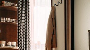 A hotel room with a wooden floor, a bed with white bedding, a brown coat hanging on a hook, a wooden sideboard with glassware, and patterned curtains.