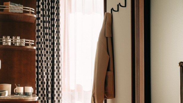 A hotel room with a wooden floor, a bed with white bedding, a brown coat hanging on a hook, a wooden sideboard with glassware, and patterned curtains.