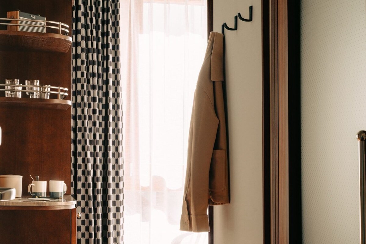 Une chambre d’hôtel avec un plancher en bois, un lit recouvert de draps blancs, un manteau marron accroché à un crochet, une commode en bois sur laquelle sont posés des verres, et des rideaux à motifs.