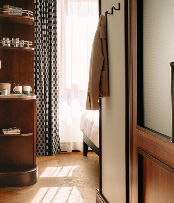 Une chambre d’hôtel avec un plancher en bois, un lit recouvert de draps blancs, un manteau marron accroché à un crochet, une commode en bois sur laquelle sont posés des verres, et des rideaux à motifs.